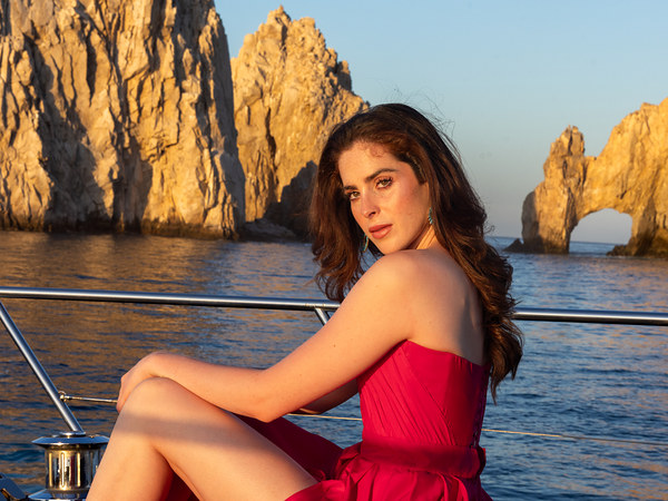 Woman in a red gown seated on a yacht at golden hour with El Arco and the Land's End rocks behind her in Cabo San Lucas
